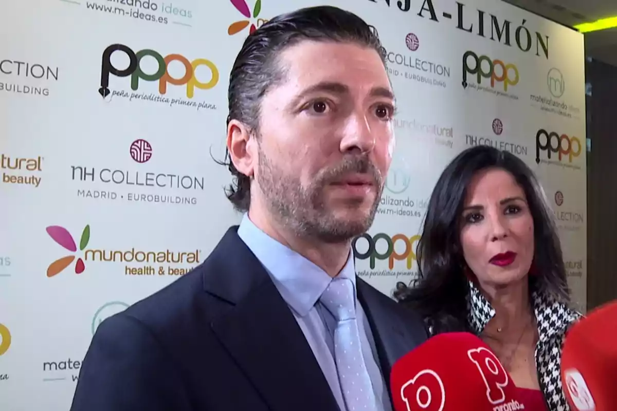 Ángel Cristoo amb vestit i corbata és entrevistat per la premsa mentre Ana Herminia amb llavis vermells l’acompanya, tots dos estan davant d’un fons amb logotips de diferents marques i mitjans. Ángel Cristoo amb vestit i corbata és entrevistat per la premsa mentre Ana Herminia amb llavis vermells l’acompanya, tots dos estan davant d’un fons amb logotips de diferents marques i mitjans.