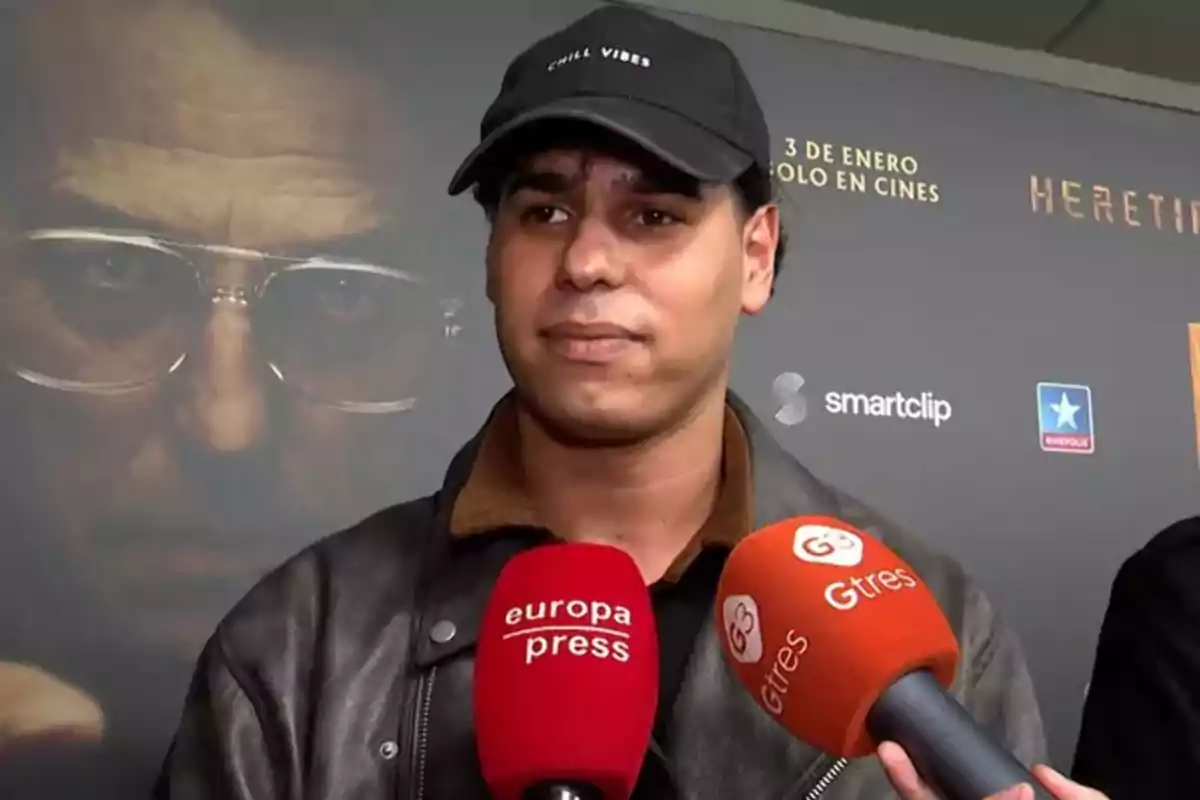 Anuar Beno amb gorra negra i jaqueta fosca és entrevistat per diversos mitjans de comunicació davant d'un cartell de cinema.