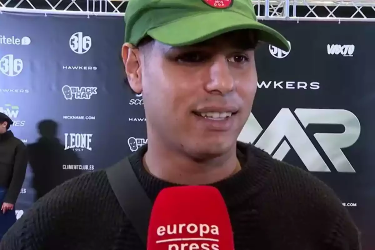 Anuar Beno amb gorra verda és entrevistat per Europa Press en un esdeveniment amb un fons de logotips de marques.