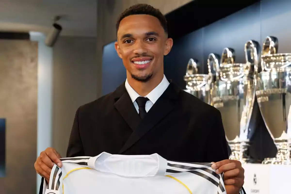 Hombre sonriente con traje sosteniendo una camiseta de fútbol blanca frente a varios trofeos plateados Hombre sonriente con traje sosteniendo una camiseta de fútbol blanca frente a varios trofeos plateados