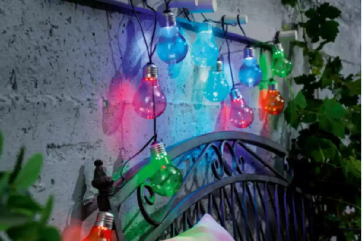 Garlanda de llums de colors enceses decorant una paret de ciment al costat d’un llit de ferro en un espai exterior amb plantes. Garlanda de llums de colors enceses decorant una paret de ciment al costat d’un llit de ferro en un espai exterior amb plantes.