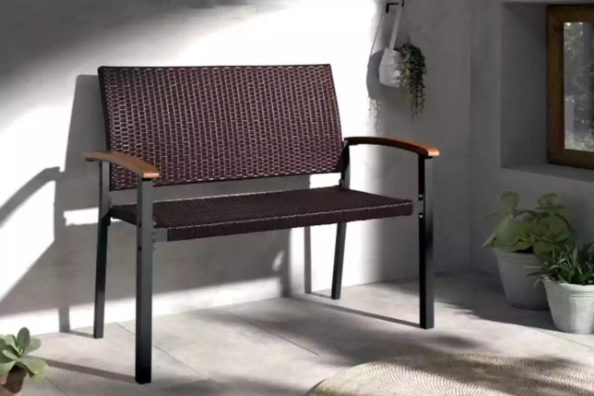 Un banc de vímet fosc amb estructura de metall i reposabraços de fusta es troba en un espai interior il·luminat per la llum natural, envoltat de plantes en testos. Un banc de vímet fosc amb estructura de metall i reposabraços de fusta es troba en un espai interior il·luminat per la llum natural, envoltat de plantes en testos.