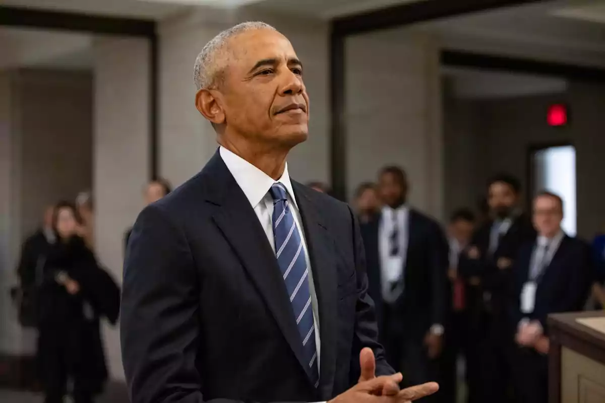 Barack Obama vestit amb un vestit fosc i corbata de ratlles, dret en un entorn formal amb diverses persones desenfocades al fons Barack Obama vestit amb un vestit fosc i corbata de ratlles, dret en un entorn formal amb diverses persones desenfocades al fons