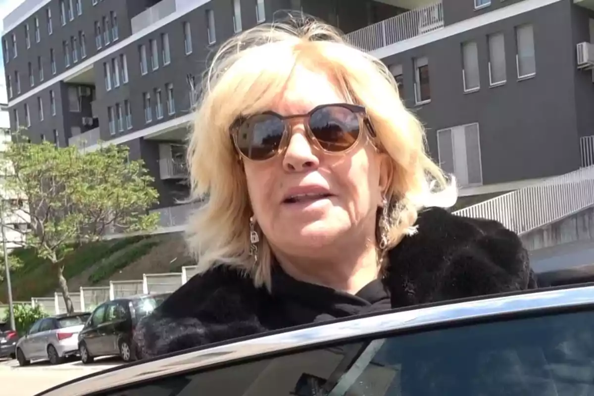 Bárbara Rey amb ulleres de sol i cabell ros està dreta al costat d'un automòbil, amb un edifici modern de fons. Bárbara Rey amb ulleres de sol i cabell ros està dreta al costat d'un automòbil, amb un edifici modern de fons.