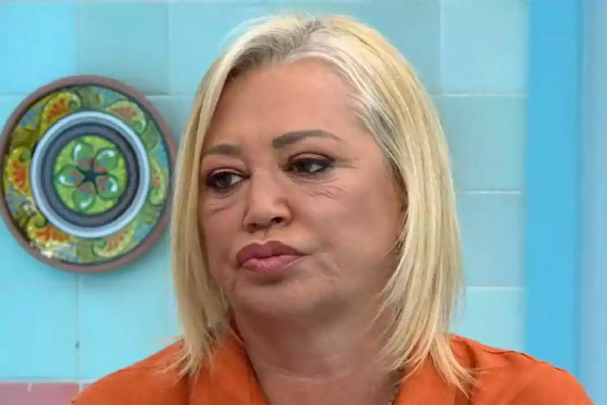 Belén Esteban de cabell ros i expressió seriosa en un entorn amb rajoles blaves i un plat decoratiu a la paret a La Família de la Tele. Belén Esteban de cabell ros i expressió seriosa en un entorn amb rajoles blaves i un plat decoratiu a la paret a La Família de la Tele.