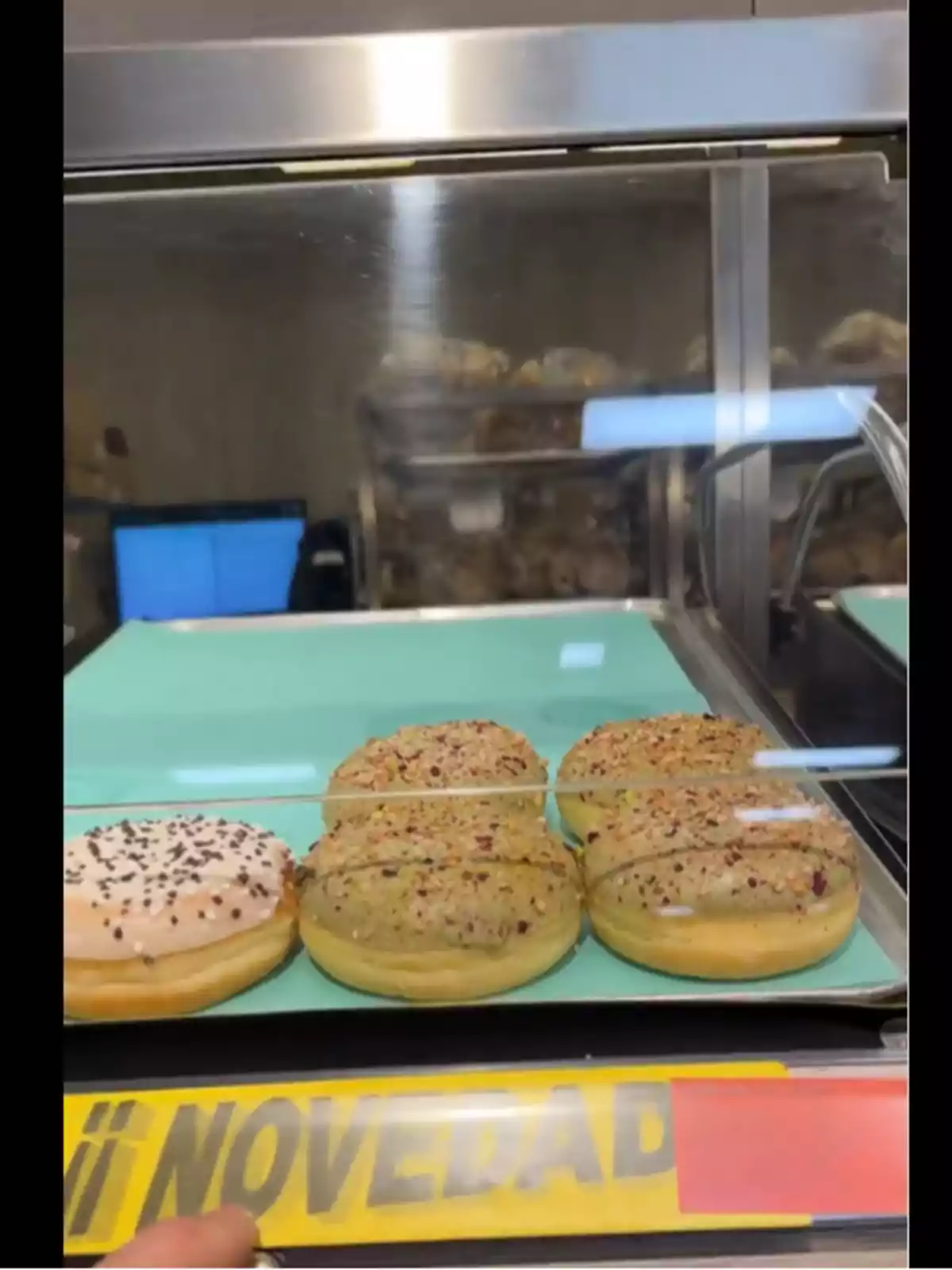 Un aparador de fleca mostra dònuts decorats amb diferents cobertures, acompanyats d'un cartell groc que indica Un aparador de fleca mostra dònuts decorats amb diferents cobertures, acompanyats d'un cartell groc que indica