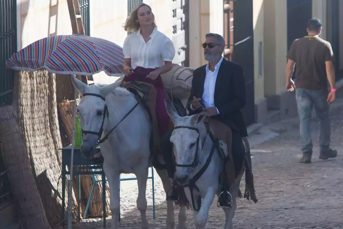 Brie Larson and George Clooney, elegantly dressed, ride donkeys on a cobblestone street while a man walks in the background and a striped umbrella is visible to one side. Brie Larson and George Clooney, elegantly dressed, ride donkeys on a cobblestone street while a man walks in the background and a striped umbrella is visible to one side.