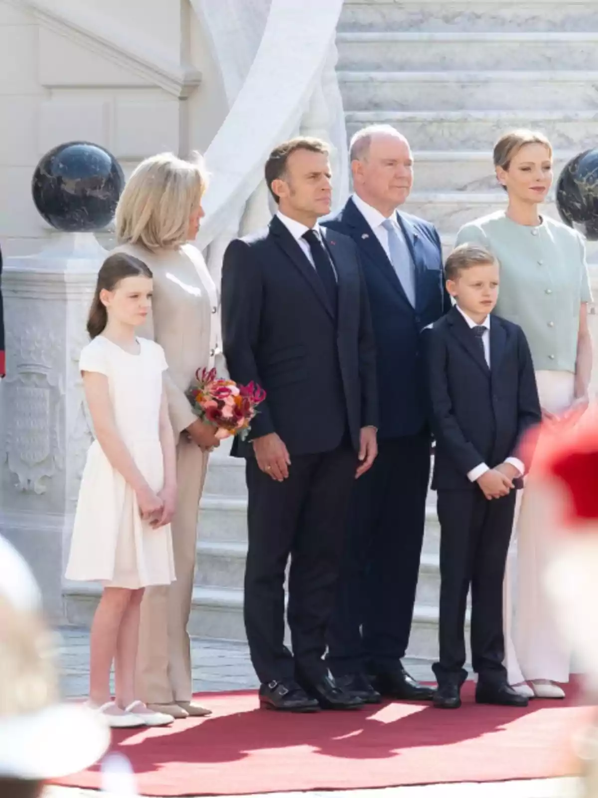 Grup de sis persones elegantment vestides posant sobre una catifa vermella davant d’una escala de marbre Grup de sis persones elegantment vestides posant sobre una catifa vermella davant d’una escala de marbre