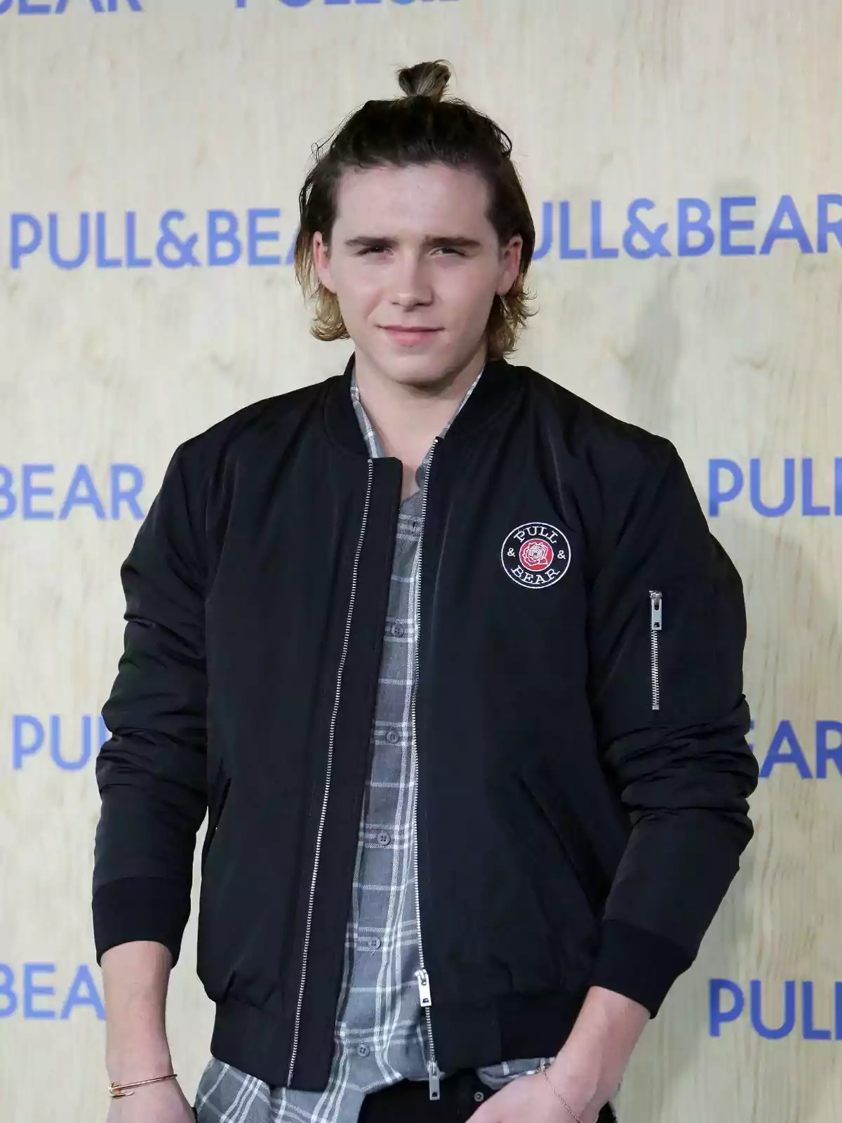Brooklyn Beckham in a black jacket and checked shirt poses in front of a background with the Pull&Bear logo. Brooklyn Beckham in a black jacket and checked shirt poses in front of a background with the Pull&Bear logo.