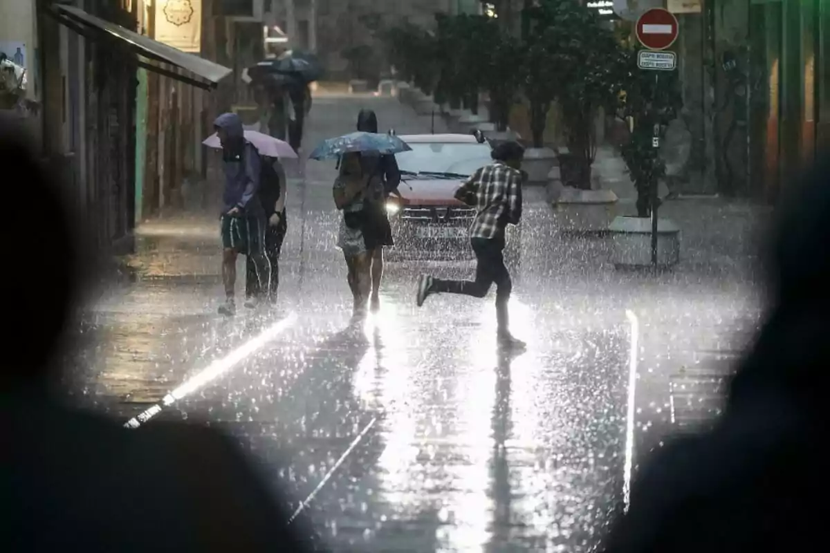 Imatge d'un carrer mentre plou amb persones corrent, altres amb paraigües i un cotxe circulant Imatge d'un carrer mentre plou amb persones corrent, altres amb paraigües i un cotxe circulant