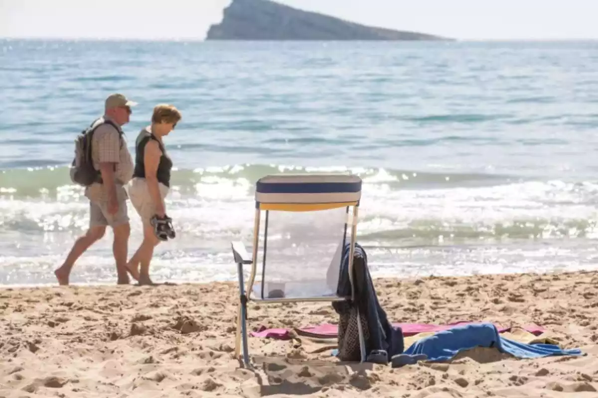 Dues persones grans passegen a la vora de la platja, amb una cadira buida a la sorra i el mar de fons