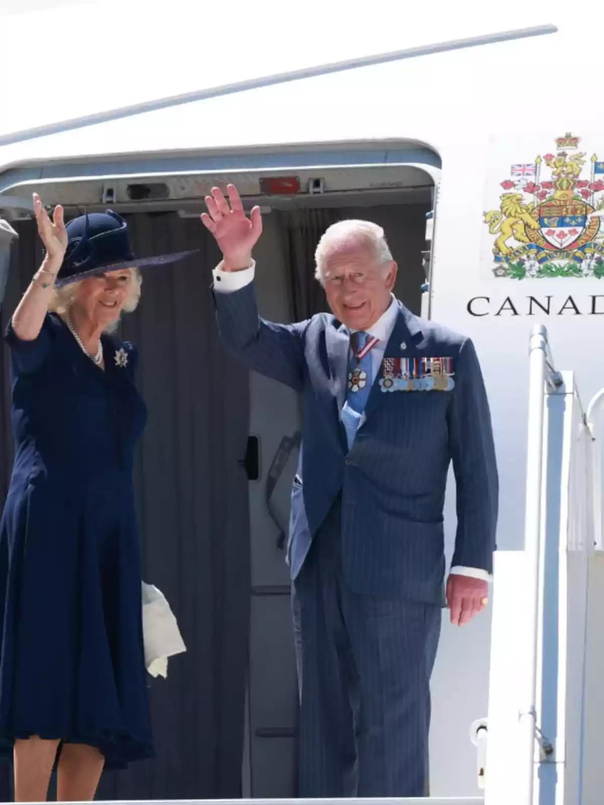 Una parella elegantament vestida saluda des de la porta d’un avió amb l’escut del Canadà visible a un costat. Una parella elegantament vestida saluda des de la porta d’un avió amb l’escut del Canadà visible a un costat.