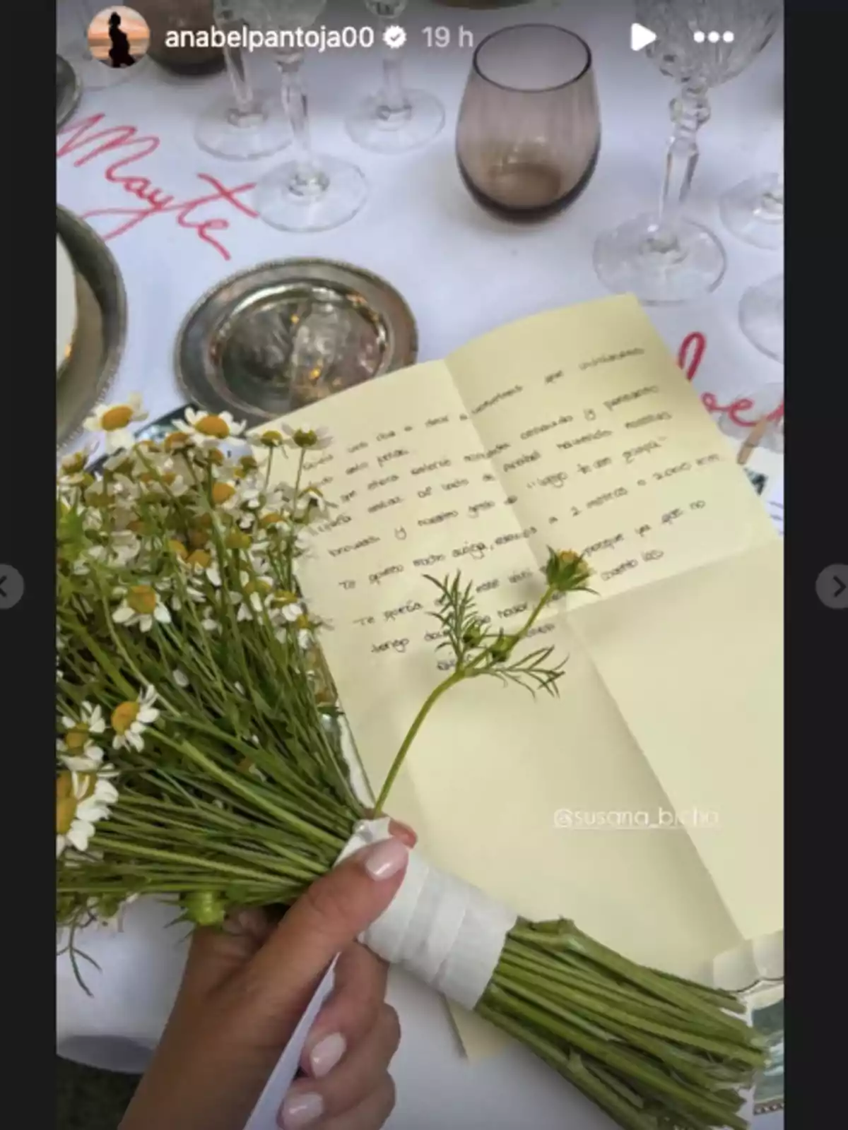 Una mano sostiene un ramo de flores pequeñas junto a una carta escrita a mano sobre una mesa elegante con copas y platos Una mano sostiene un ramo de flores pequeñas junto a una carta escrita a mano sobre una mesa elegante con copas y platos