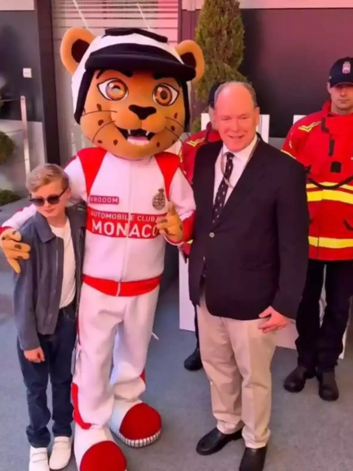 Jacques con gafas de sol, una persona disfrazada de tigre con un uniforme del Automobile Club de Mónaco y Alberto de Mónaco de traje posan juntos mientras dos personas con uniformes rojos están de pie detrás de ellos. Jacques con gafas de sol, una persona disfrazada de tigre con un uniforme del Automobile Club de Mónaco y Alberto de Mónaco de traje posan juntos mientras dos personas con uniformes rojos están de pie detrás de ellos.