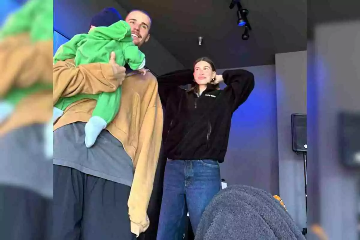 Justin Bieber sosté un nadó vestit de verd mentre Hailey Bieber al seu costat somriu i es toca els cabells en una habitació il·luminada amb llums blaves. Justin Bieber sosté un nadó vestit de verd mentre Hailey Bieber al seu costat somriu i es toca els cabells en una habitació il·luminada amb llums blaves.