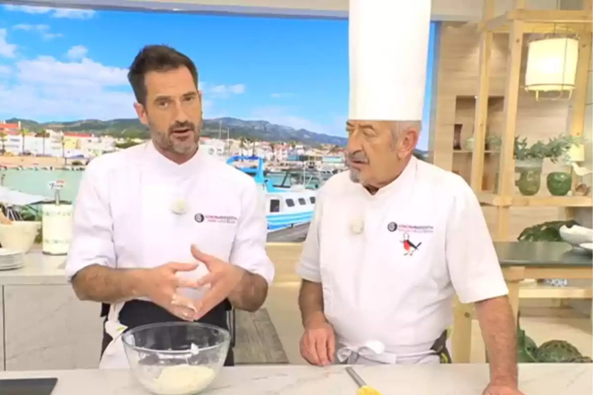 Karlos i Joseba Arguiñano amb uniforme blanc conversen en una cuina moderna mentre Joseba barreja ingredients en un bol de vidre i Karlos porta un barret alt de xef. Karlos i Joseba Arguiñano amb uniforme blanc conversen en una cuina moderna mentre Joseba barreja ingredients en un bol de vidre i Karlos porta un barret alt de xef.