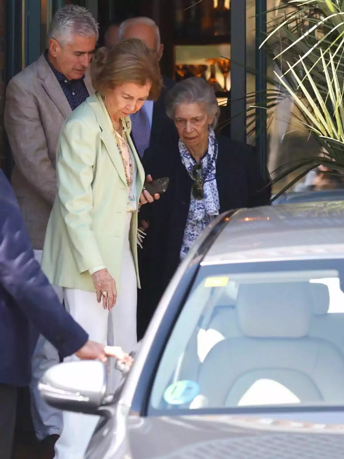 A group of elderly people exit a building and walk towards a car parked nearby. A group of elderly people exit a building and walk towards a car parked nearby.