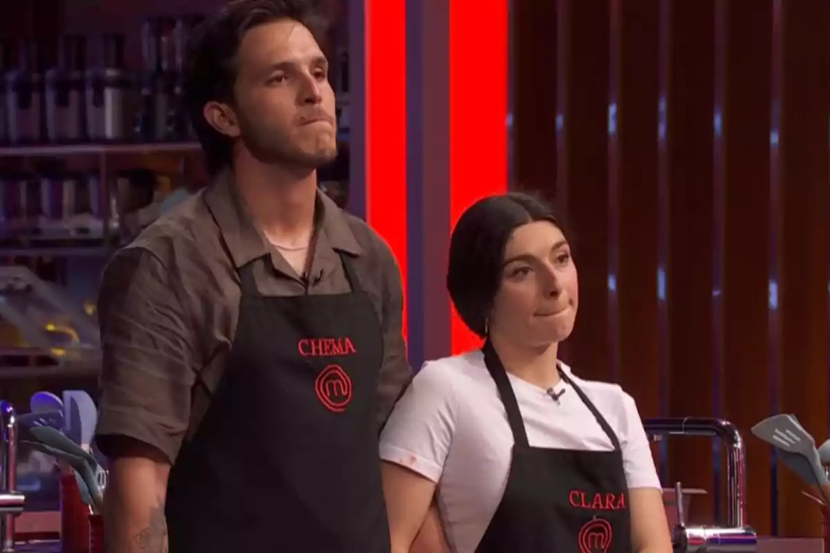 Clara i Chema, concursants de 'Masterchef' amb davantals negres i expressió seriosa en una cuina il·luminada amb llums vermelles. Clara i Chema, concursants de 'Masterchef' amb davantals negres i expressió seriosa en una cuina il·luminada amb llums vermelles.