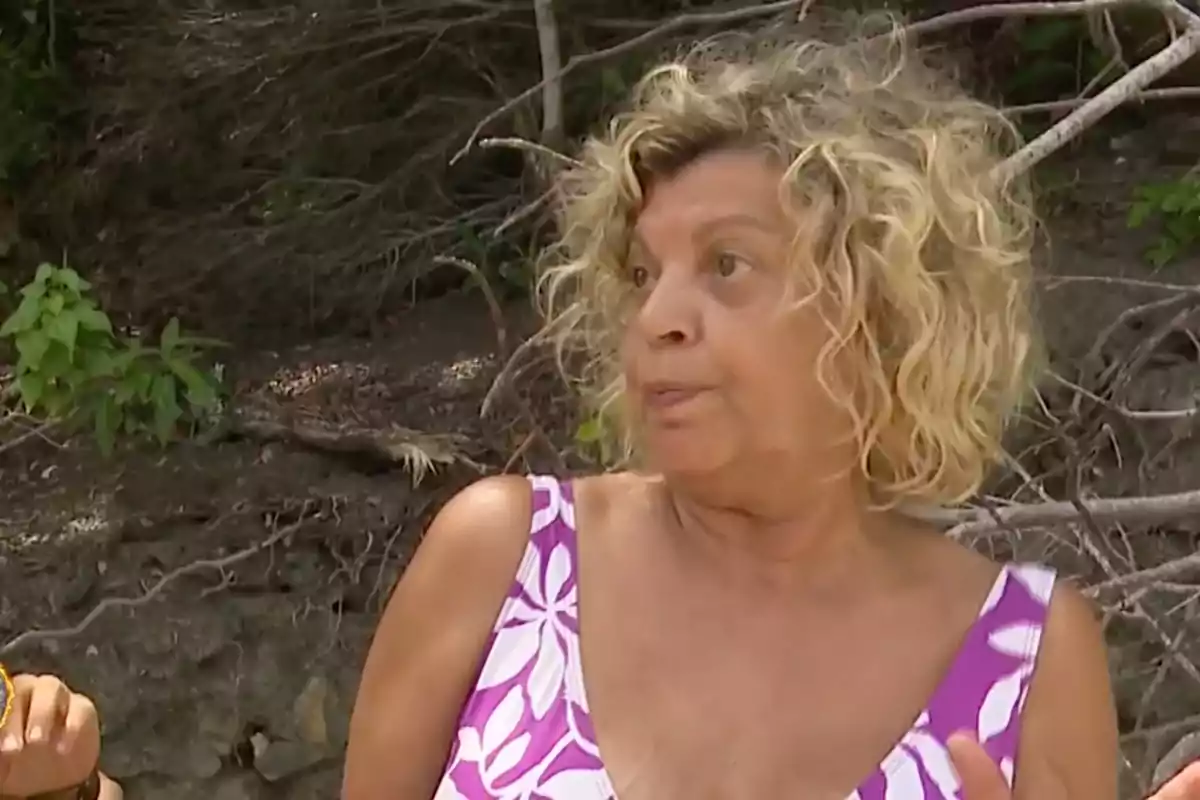 Terelu Campos con cabello rizado rubio y vestido con estampado floral púrpura está al aire libre con un fondo de ramas y vegetación. Terelu Campos con cabello rizado rubio y vestido con estampado floral púrpura está al aire libre con un fondo de ramas y vegetación.