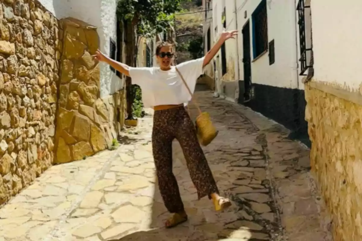 Nuria Roca con gafas de sol y una bolsa de mimbre posa alegremente en una calle estrecha y empedrada, rodeada de edificios de piedra y paredes blancas. Nuria Roca con gafas de sol y una bolsa de mimbre posa alegremente en una calle estrecha y empedrada, rodeada de edificios de piedra y paredes blancas.