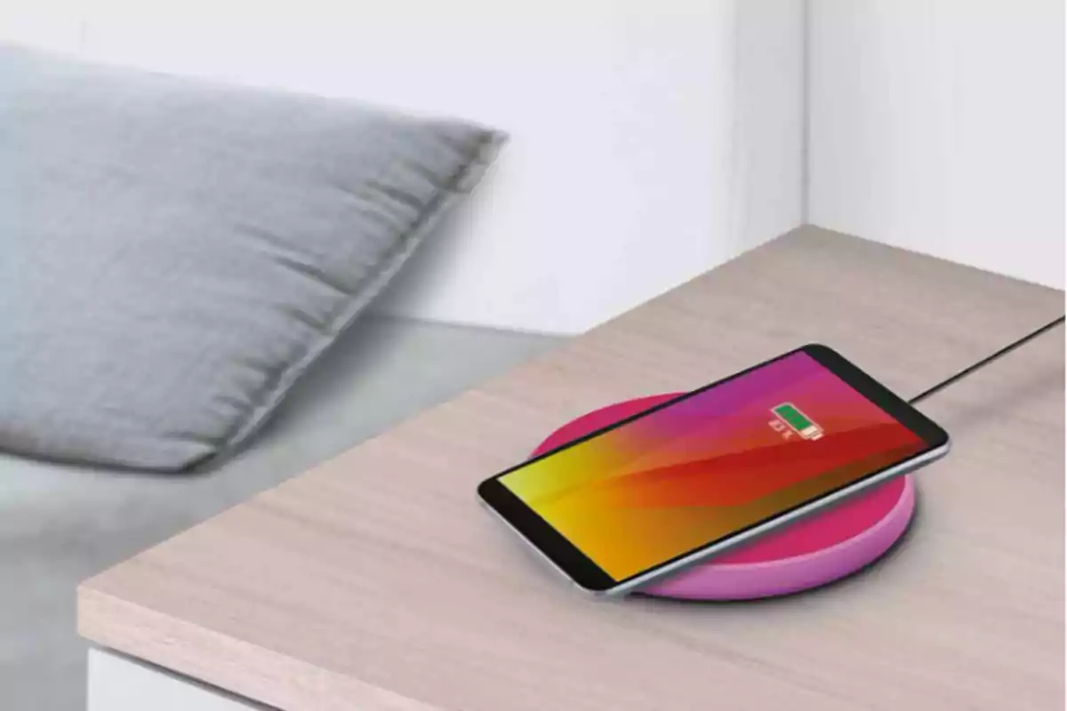 Telèfon intel·ligent carregant sense fil sobre una base rosa en una taula de fusta al costat d'un coixí gris. Telèfon intel·ligent carregant sense fil sobre una base rosa en una taula de fusta al costat d'un coixí gris.