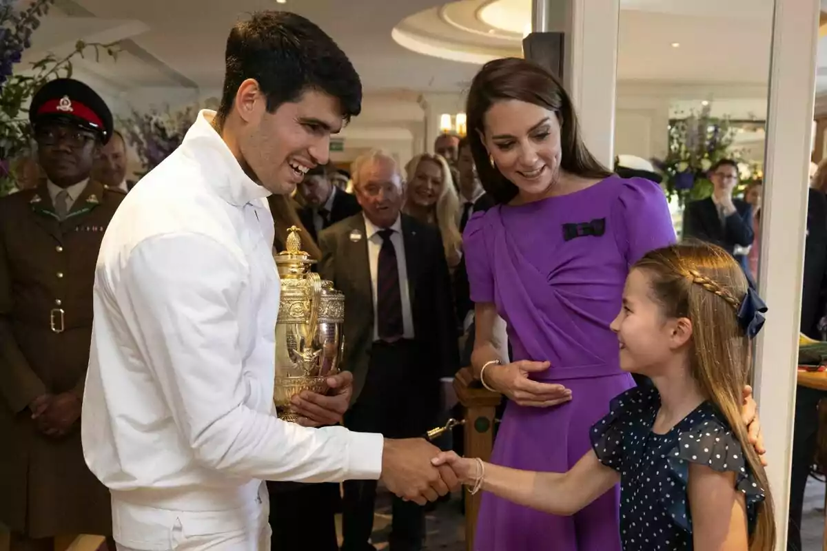 Carlos Alcaraz vestit de blanc sosté un trofeu mentre estreny la mà de Charlotte, acompanyat per Kate Middleton vestida de morat i diverses persones al fons. Carlos Alcaraz vestit de blanc sosté un trofeu mentre estreny la mà de Charlotte, acompanyat per Kate Middleton vestida de morat i diverses persones al fons.