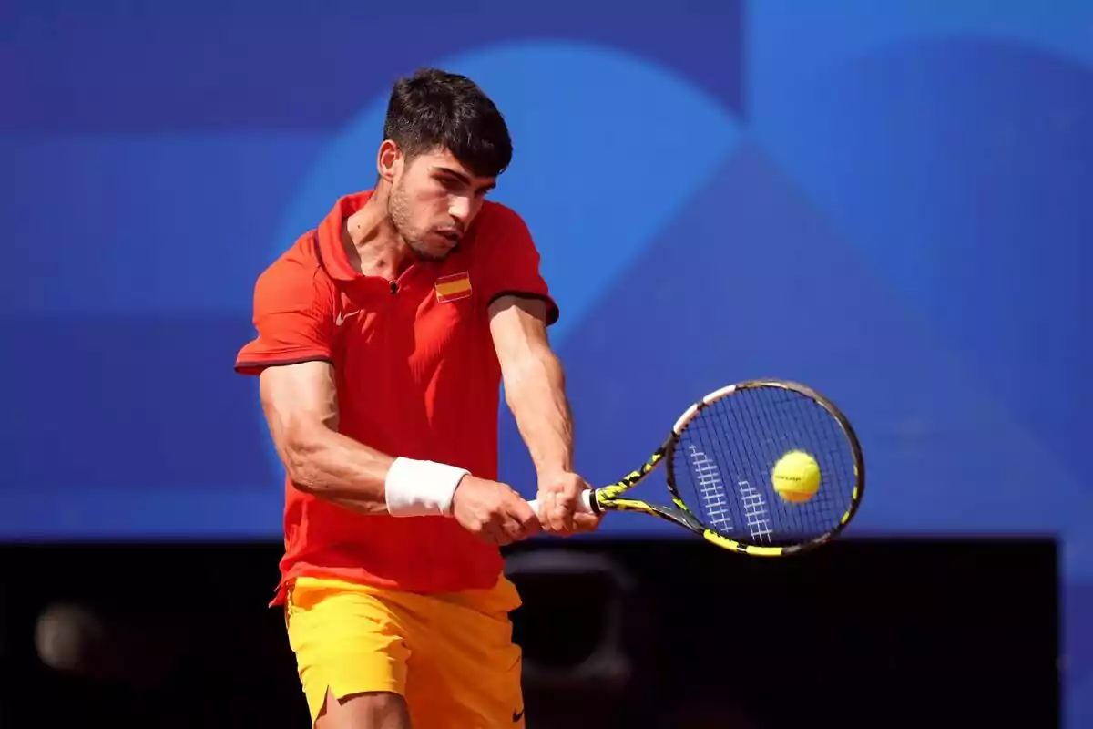 Carlos Alcaraz colpejant la pilota amb una raqueta durant un partit, vestint una samarreta vermella i pantalons curts grocs. Carlos Alcaraz colpejant la pilota amb una raqueta durant un partit, vestint una samarreta vermella i pantalons curts grocs.