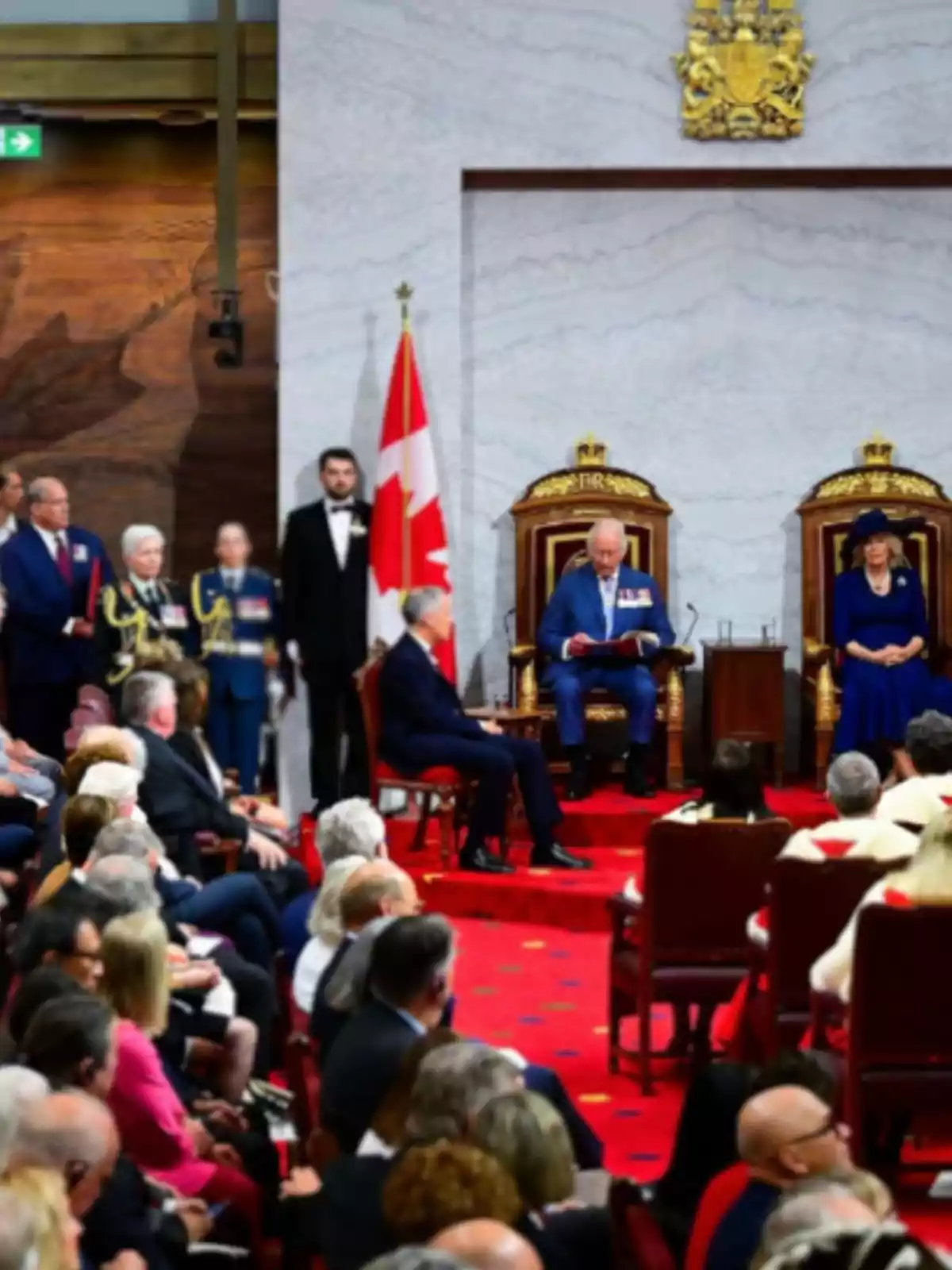Cerimònia oficial en una sala amb catifa vermella, dues persones assegudes en trons daurats, una bandera del Canadà i públic observant. Cerimònia oficial en una sala amb catifa vermella, dues persones assegudes en trons daurats, una bandera del Canadà i públic observant.