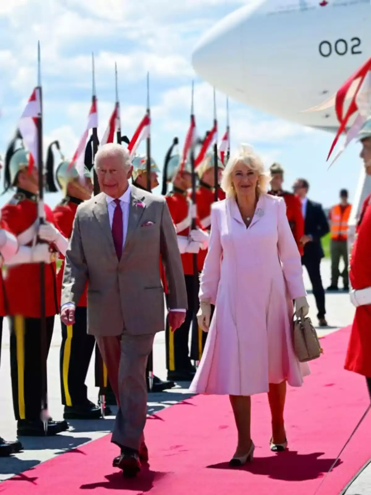 Una parella elegantament vestida camina per una catifa vermella mentre soldats amb uniformes de gala i llances formen una guàrdia d’honor al costat d’un avió blanc. Una parella elegantament vestida camina per una catifa vermella mentre soldats amb uniformes de gala i llances formen una guàrdia d’honor al costat d’un avió blanc.