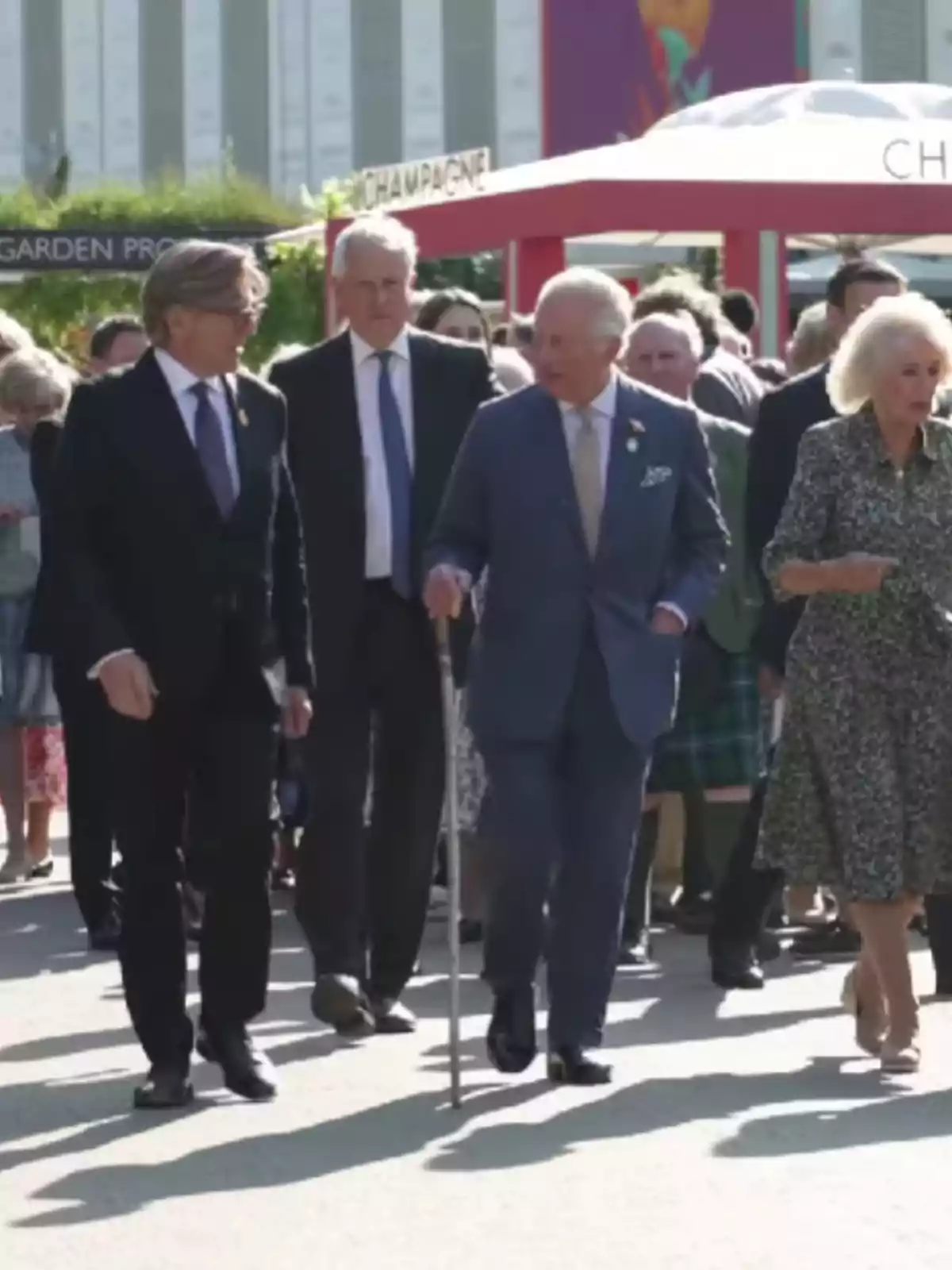 Un grup de persones caminant a l'aire lliure en un esdeveniment, amb un home gran usant un bastó i una dona amb un vestit estampat. Un grup de persones caminant a l'aire lliure en un esdeveniment, amb un home gran usant un bastó i una dona amb un vestit estampat.