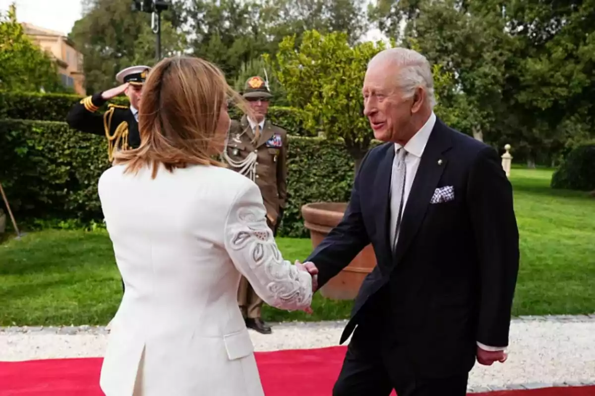 Carlos III in a dark suit shakes hands with Giorgia Meloni in a white jacket at an outdoor event, while two uniformed officers watch in the background. Carlos III in a dark suit shakes hands with Giorgia Meloni in a white jacket at an outdoor event, while two uniformed officers watch in the background.