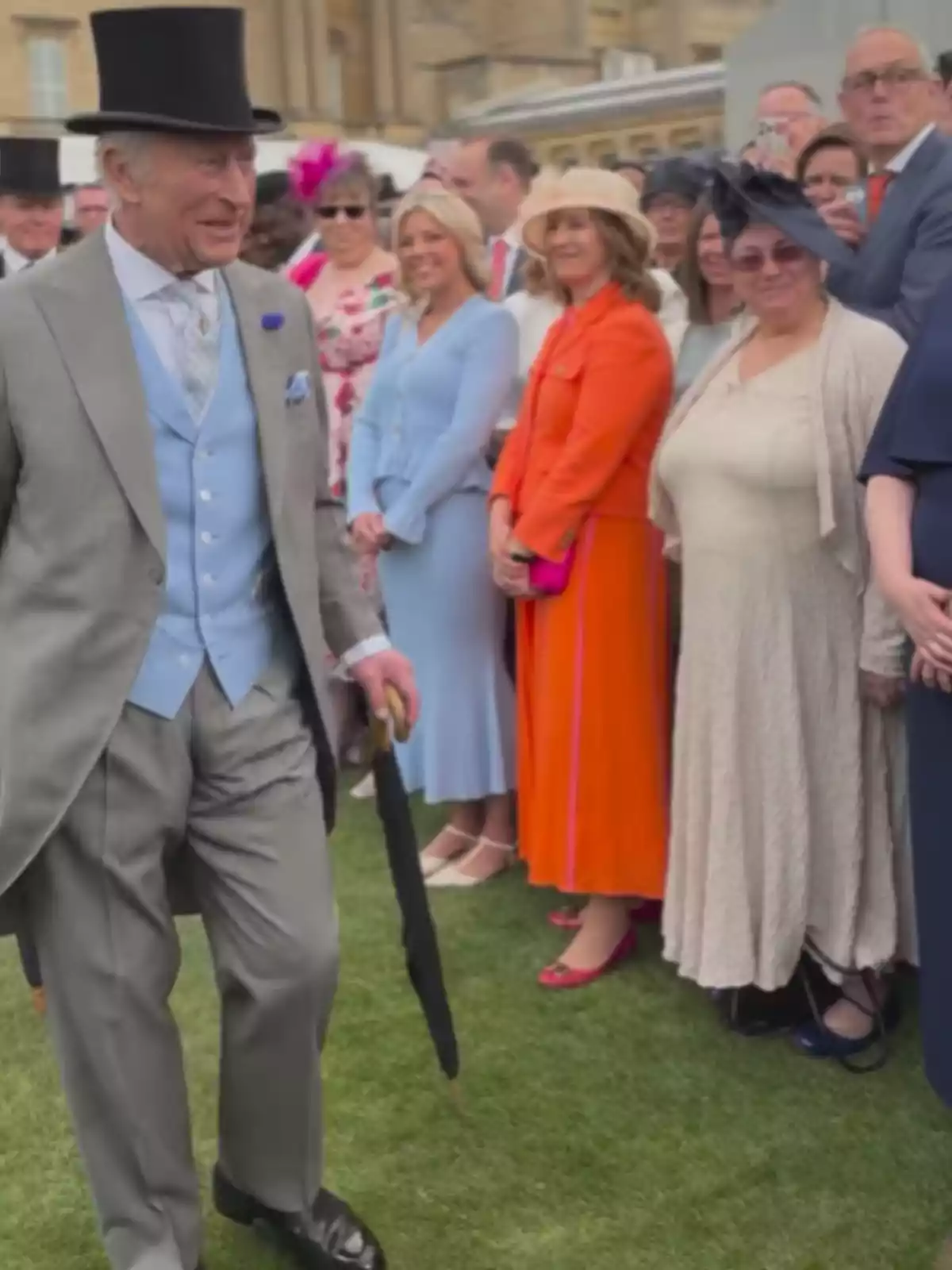An elderly man in a top hat and gray suit walks on grass while holding an umbrella, surrounded by elegantly dressed people. An elderly man in a top hat and gray suit walks on grass while holding an umbrella, surrounded by elegantly dressed people.