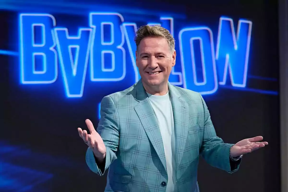 Carlos Latre wearing a green checkered jacket and light blue shirt smiles with open arms in front of a lighted sign that reads 