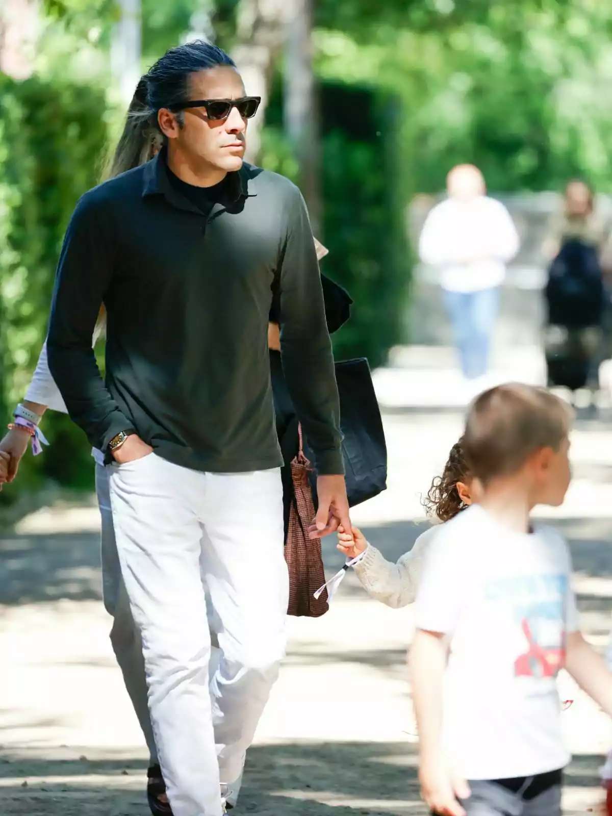 Carlos Torretta wearing sunglasses and a dark green long-sleeved shirt walks outdoors while holding the hand of a little boy. Carlos Torretta wearing sunglasses and a dark green long-sleeved shirt walks outdoors while holding the hand of a little boy.
