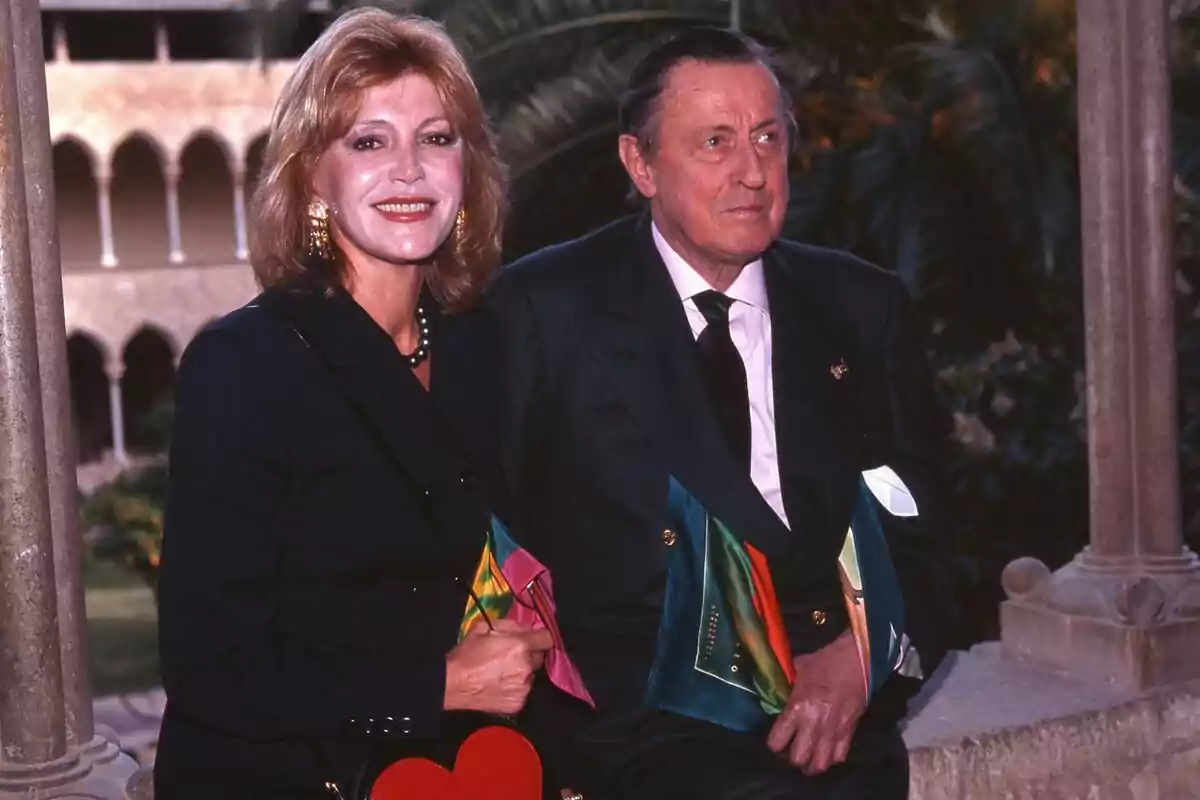 Carmen Thyssen and Heinrich Thyssen stand together outdoors, both elegantly dressed, while the woman carries a red heart-shaped bag.