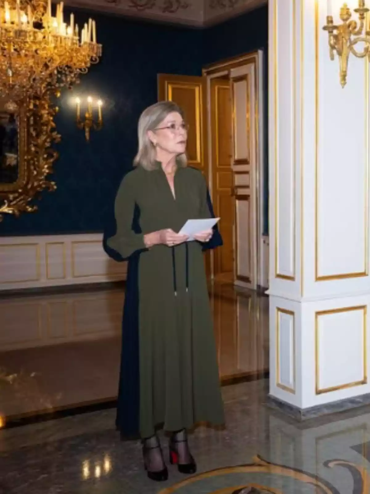 Dona de cabells canosos i vestit llarg verd fosc sosté unes fulles mentre està dreta en una elegant sala decorada amb canelobres daurats i parets blanques amb detalls daurats. Dona de cabells canosos i vestit llarg verd fosc sosté unes fulles mentre està dreta en una elegant sala decorada amb canelobres daurats i parets blanques amb detalls daurats.