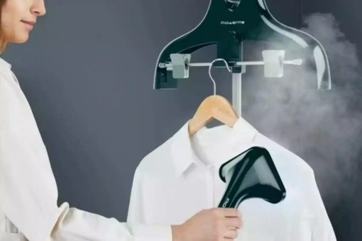 Persona utilitzant un vaporitzador de roba per treure arrugues d’una camisa blanca penjada en una perxa Persona utilitzant un vaporitzador de roba per treure arrugues d’una camisa blanca penjada en una perxa