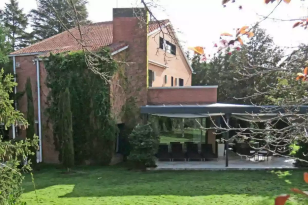 Casa de maó amb teulada a dues aigües envoltada de vegetació i un jardí ampli amb terrassa coberta Casa de maó amb teulada a dues aigües envoltada de vegetació i un jardí ampli amb terrassa coberta