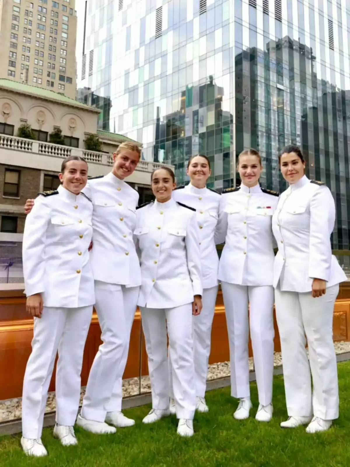 Six women dressed in formal white uniforms pose smiling outdoors in front of modern and old buildings. Six women dressed in formal white uniforms pose smiling outdoors in front of modern and old buildings.