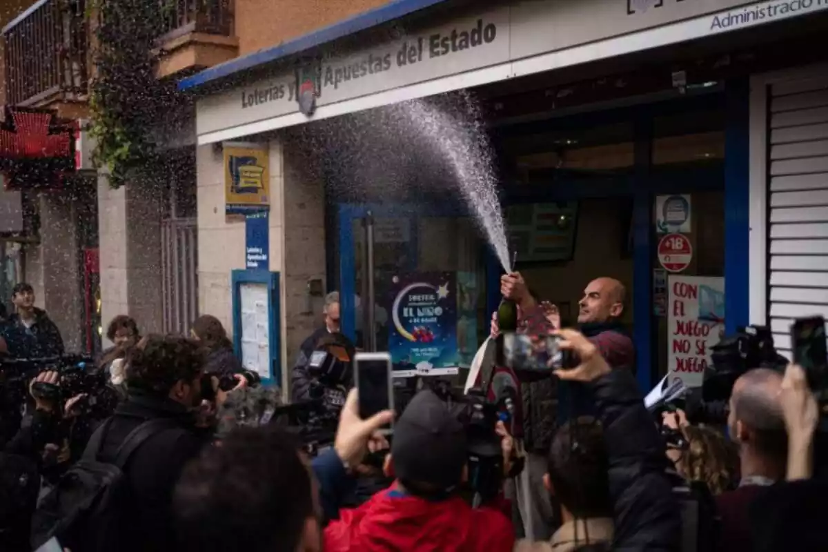 A man from the lottery department celebrates his Christmas lottery prize with a bottle of cava and people around him.