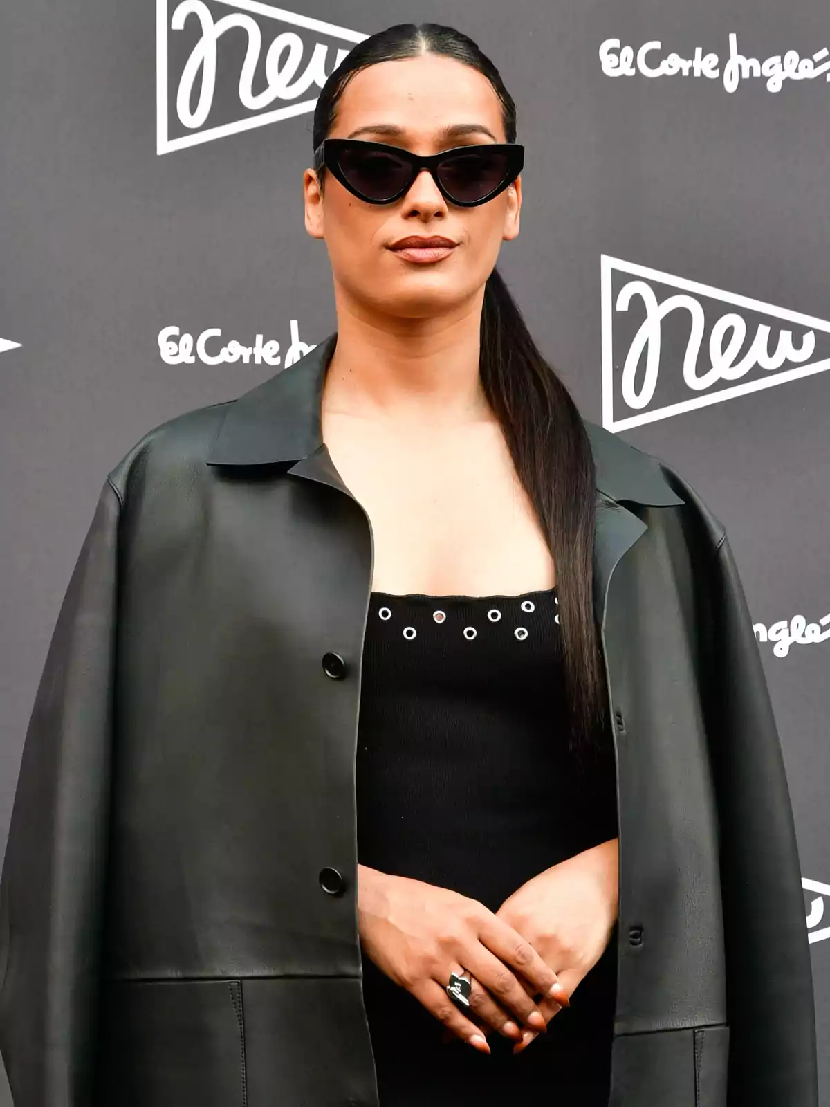 Chanel amb ulleres de sol i cabell llarg recollit porta un abric negre sobre un vestit negre amb detalls metàl·lics, posant davant d'un fons amb logotips d'El Corte Inglés. Chanel amb ulleres de sol i cabell llarg recollit porta un abric negre sobre un vestit negre amb detalls metàl·lics, posant davant d'un fons amb logotips d'El Corte Inglés.