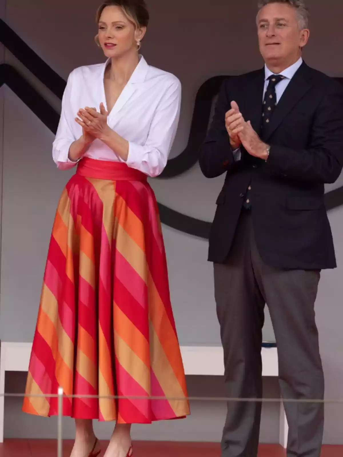 Una dona amb brusa blanca i faldilla de ratlles vermelles i taronges està dreta al costat d'un home amb vestit fosc, tots dos aplaudint. Una dona amb brusa blanca i faldilla de ratlles vermelles i taronges està dreta al costat d'un home amb vestit fosc, tots dos aplaudint.