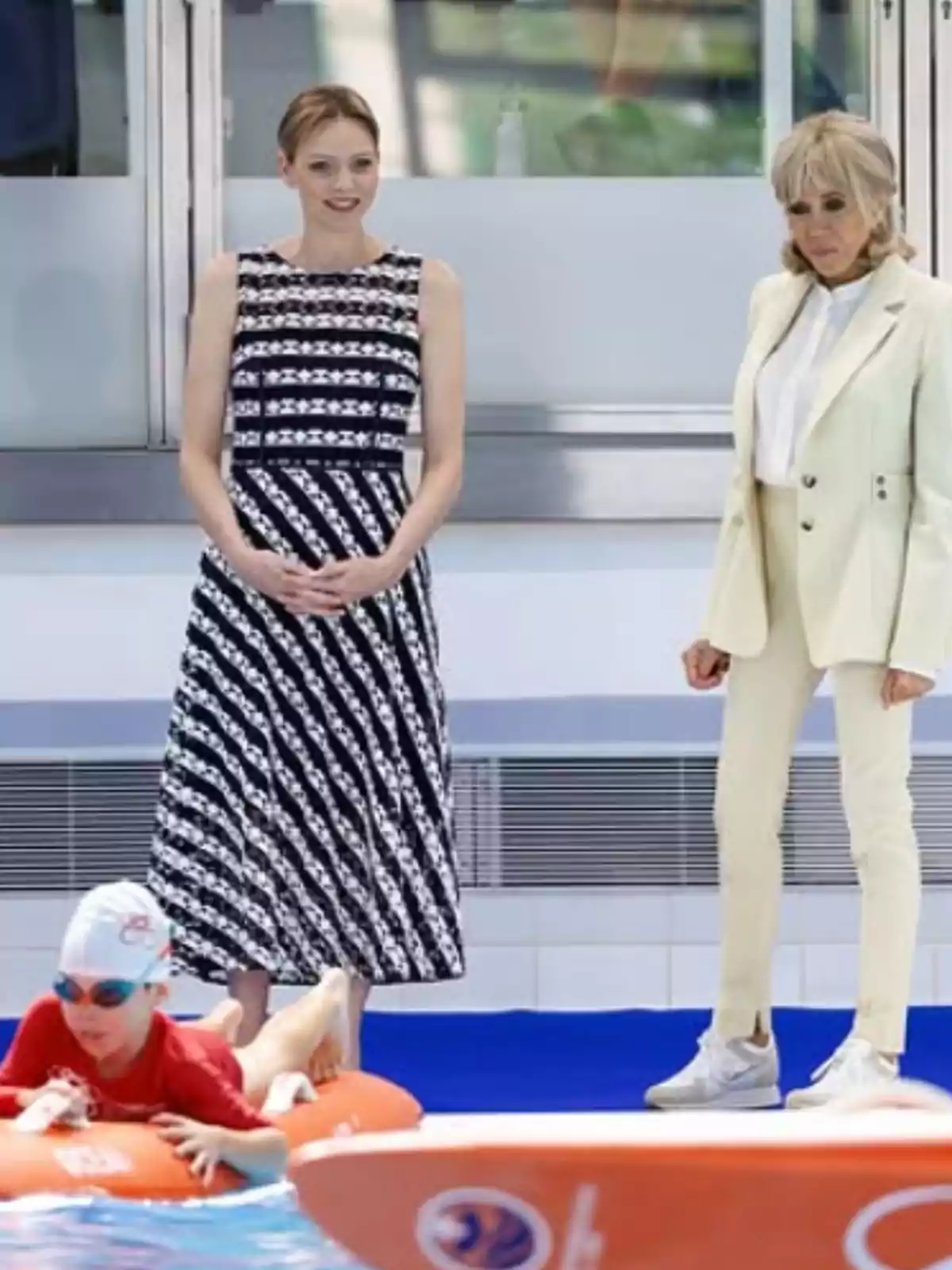 Two women watch a boy wearing a swim cap and a red jersey floating on a board in a pool. Two women watch a boy wearing a swim cap and a red jersey floating on a board in a pool.