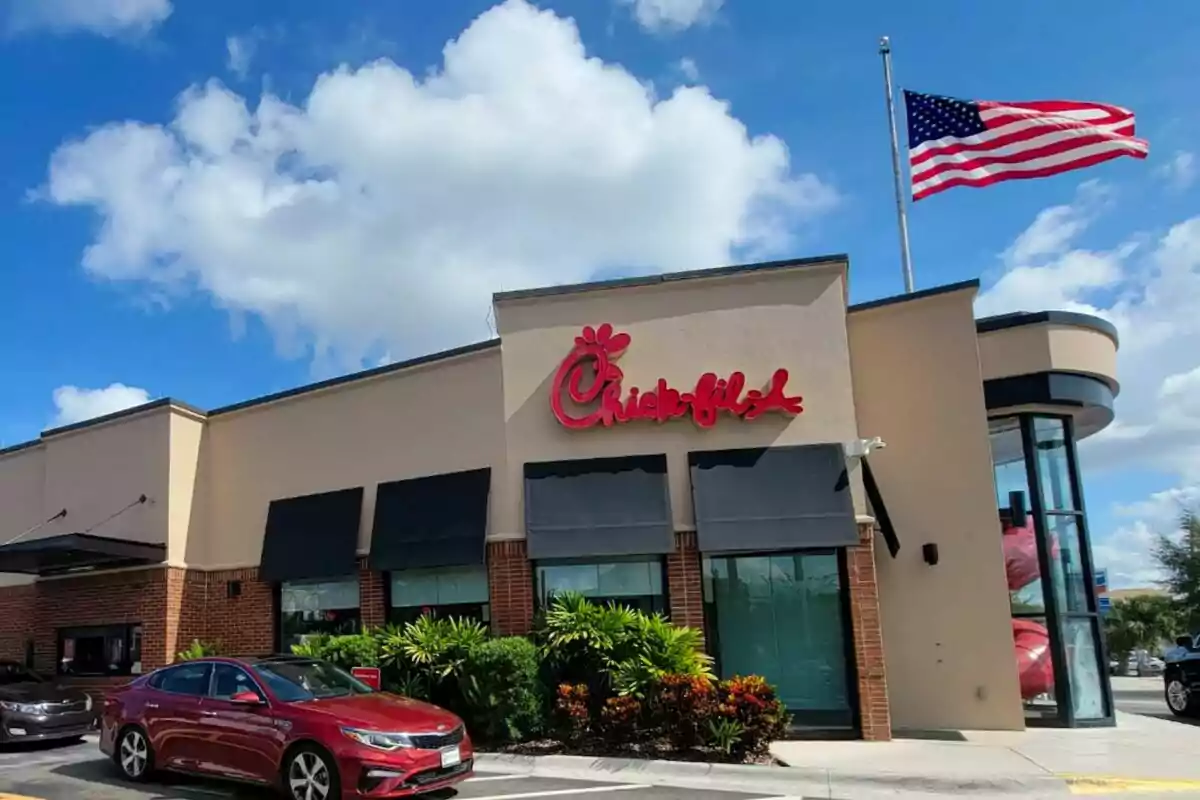 Un restaurante de comida rápida, que es Chick-fil-A, con un letrero rojo en la fachada y una bandera estadounidense ondeando en el cielo. Un restaurante de comida rápida, que es Chick-fil-A, con un letrero rojo en la fachada y una bandera estadounidense ondeando en el cielo.