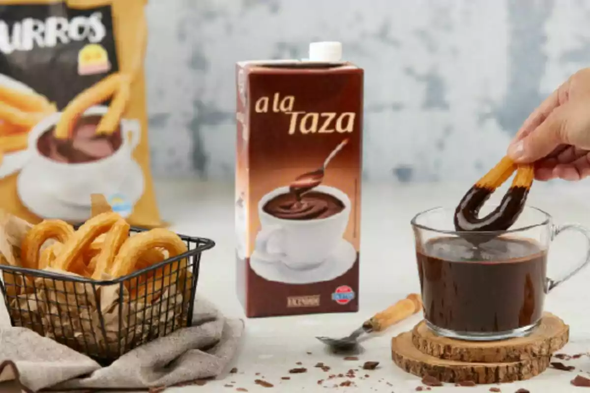 Una mà submergeix un xurro en una tassa de xocolata calenta al costat d’una capsa de xocolata a la tassa i una cistella amb xurros sobre una taula. Una mà submergeix un xurro en una tassa de xocolata calenta al costat d’una capsa de xocolata a la tassa i una cistella amb xurros sobre una taula.