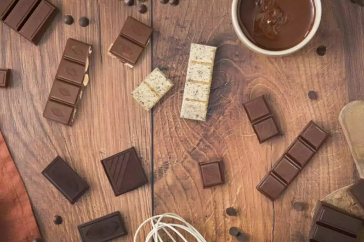 Tauleta de xocolata i trossos de xocolata de diferents tipus sobre una taula de fusta al costat d’un recipient amb xocolata desfeta i utensilis de rebosteria Tauleta de xocolata i trossos de xocolata de diferents tipus sobre una taula de fusta al costat d’un recipient amb xocolata desfeta i utensilis de rebosteria
