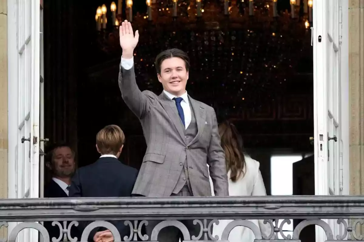 Christian de Dinamarca con traje gris y corbata azul saluda desde un balcón mientras otras personas están de espaldas. Christian de Dinamarca con traje gris y corbata azul saluda desde un balcón mientras otras personas están de espaldas.