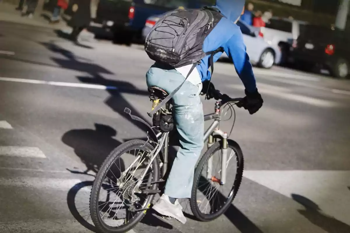 Persona amb dessuadora blava i motxilla negra muntant una bicicleta en un carrer concorregut.