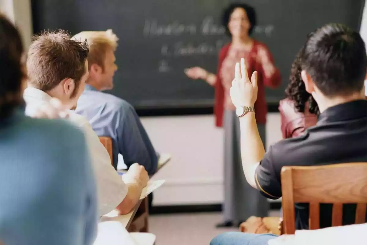 Classe universitària amb la professora a la pissarra i un estudiant d'esquena aixecant la mà