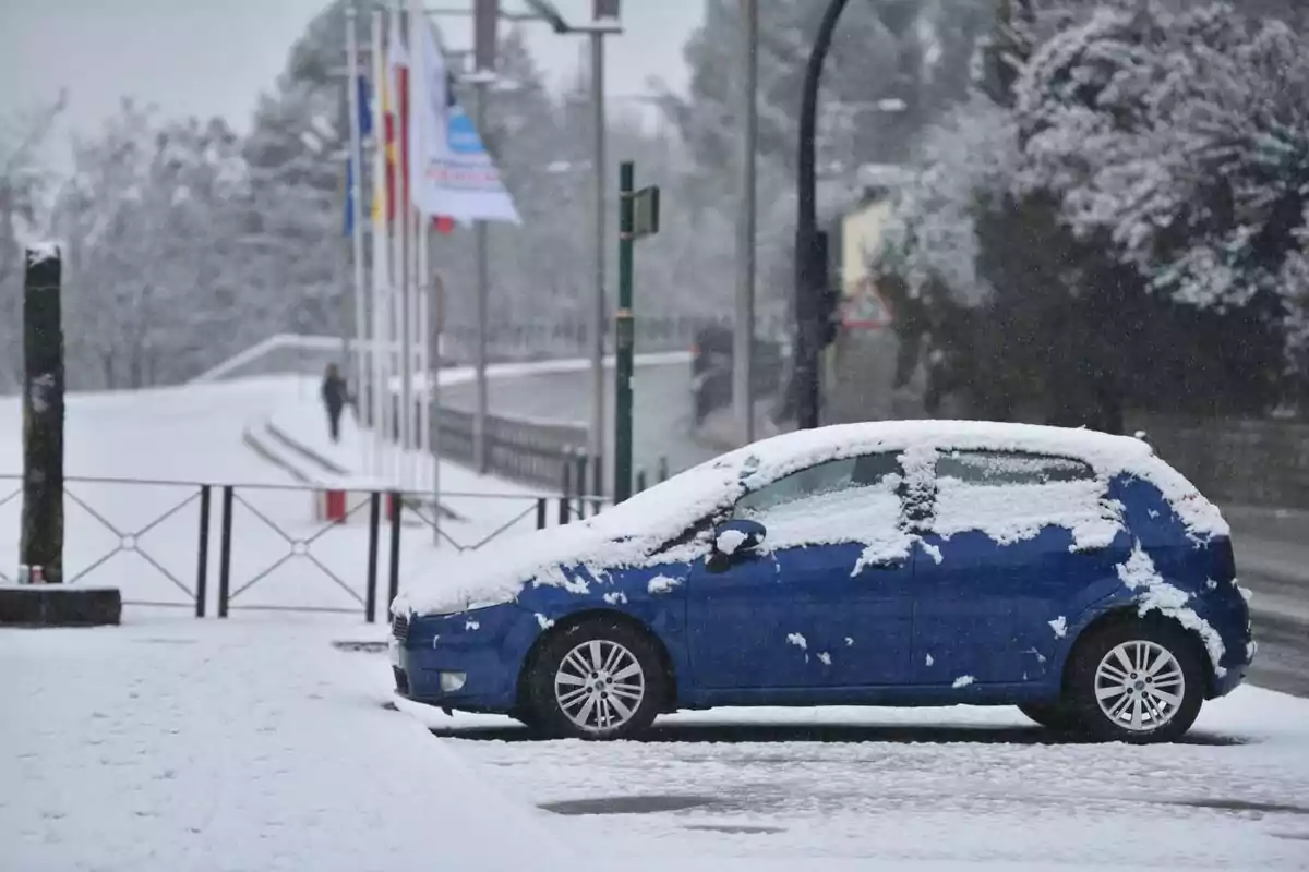 Un coche azul cubierto de nieve estacionado junto a una carretera nevada con banderas al fondo.